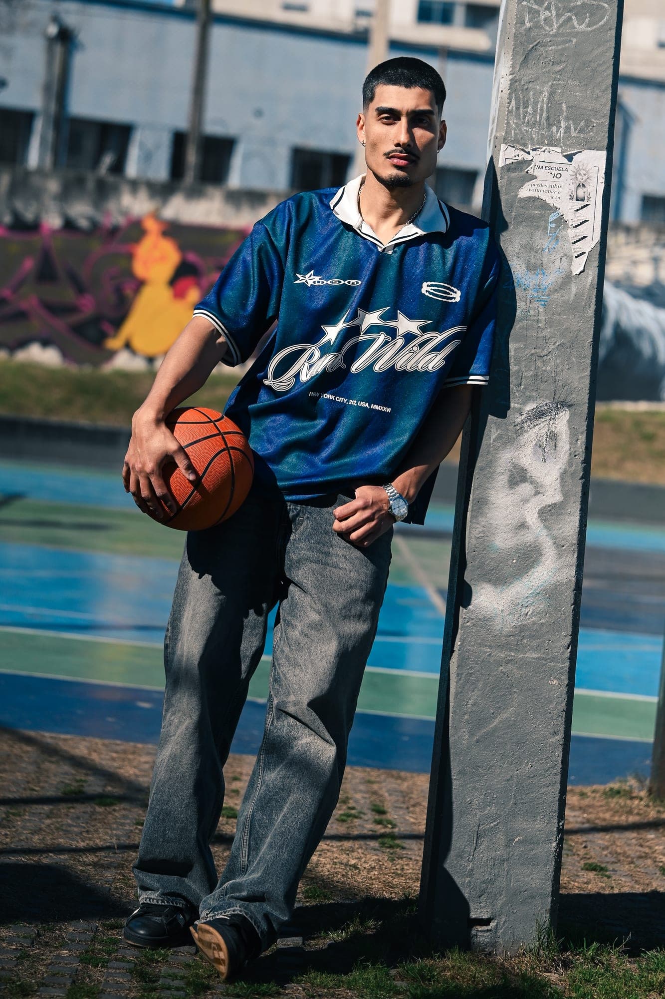 Retrato de cuerpo entero apoyado en un poste de una cancha de básquet, con la pelota en la mano