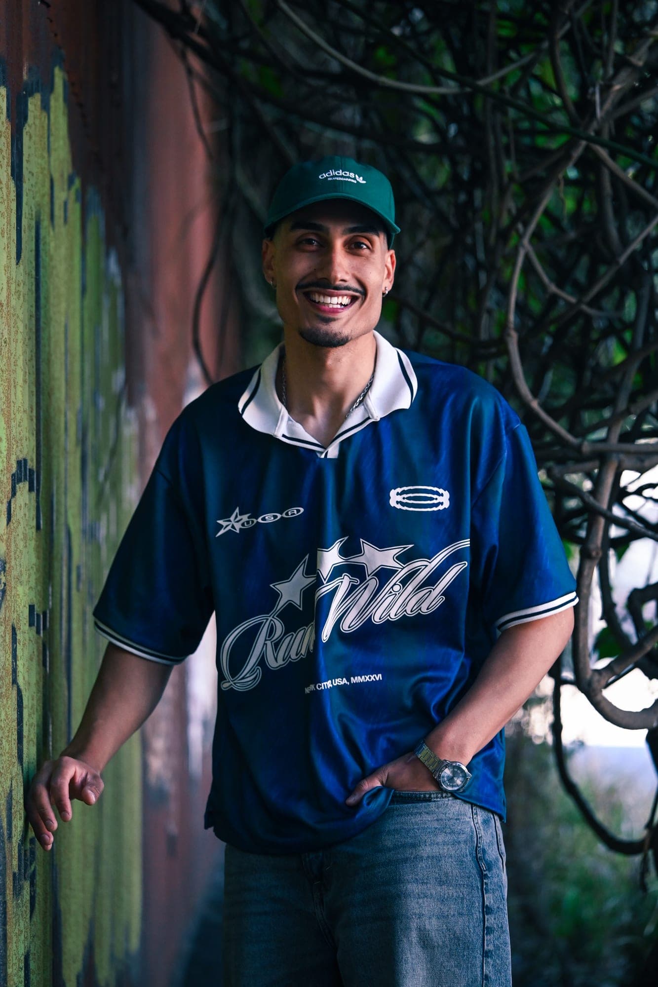 Retrato sonriendo con gorra, apoyado en una pared con graffiti bajo la vegetación